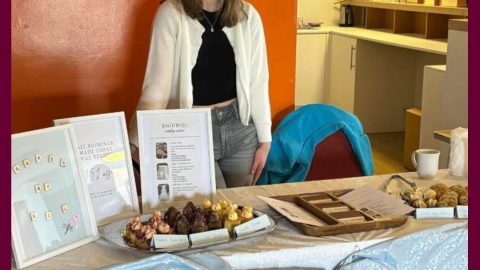 An image of a smiling woman standing behind a table with baked treats on it.